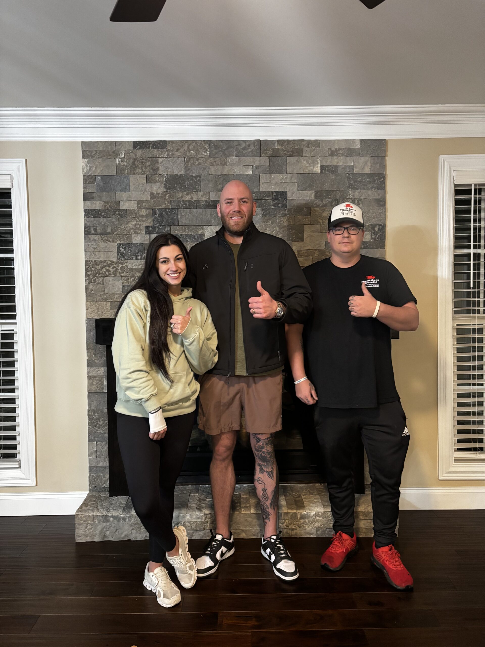 Three people stand indoors in front of a stone fireplace, smiling and giving thumbs up. The group includes one woman and two men in casual clothing.