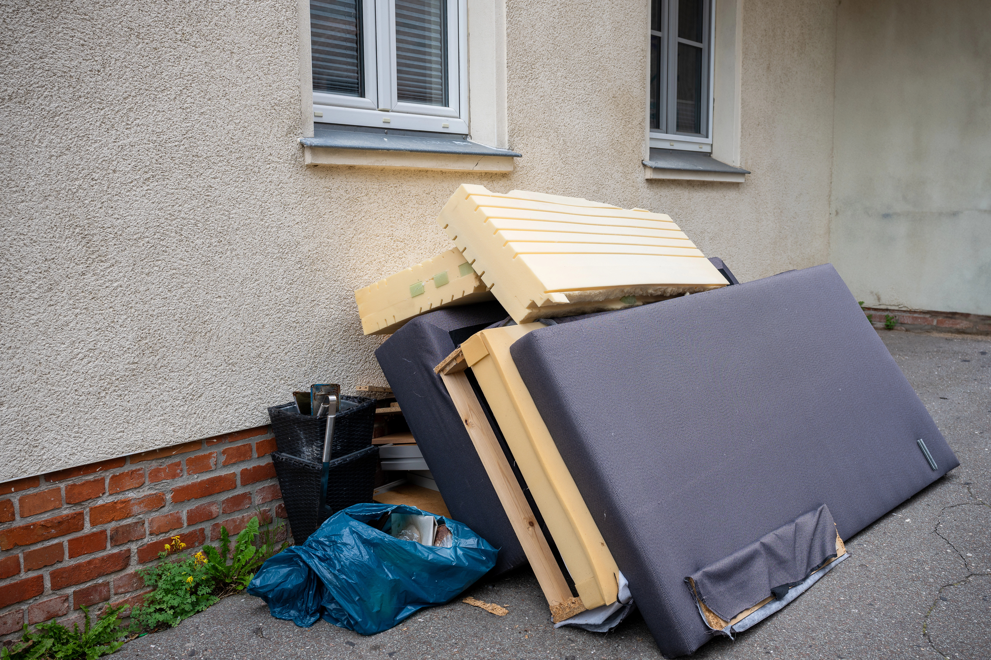 Disassembled furniture, a mattress, and bags of trash are piled against the exterior wall of a building beneath two windows.