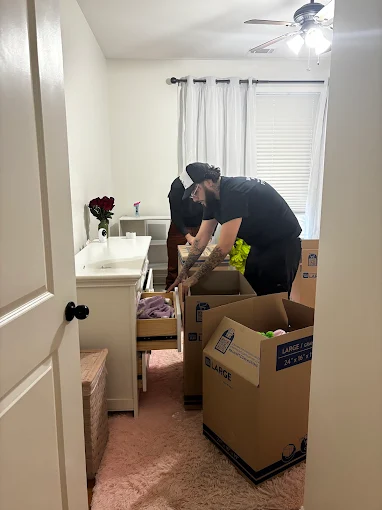 A person packs items from a dresser into cardboard boxes in a small, carpeted room with a ceiling fan and closed curtains.