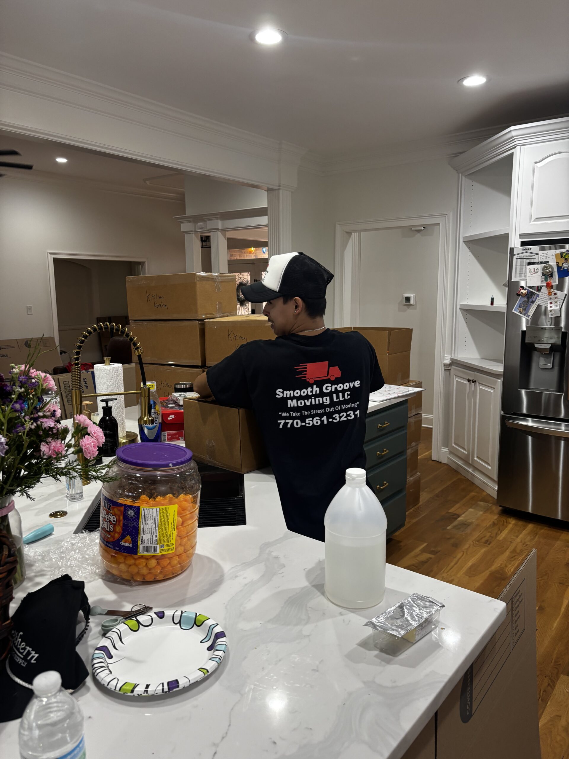 A person wearing a Smooth Groove Moving LLC shirt stands in a kitchen near a stack of cardboard boxes on the counter. Nearby are snacks, a gallon jug, flowers, and kitchen items.